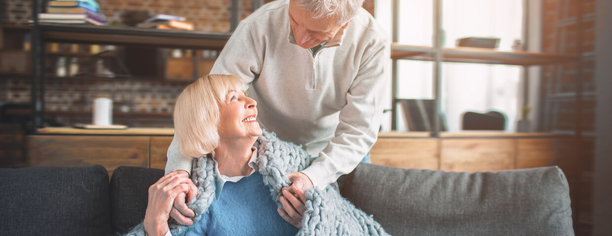 elder man putting a blanket on elder woman while she sits on the couch